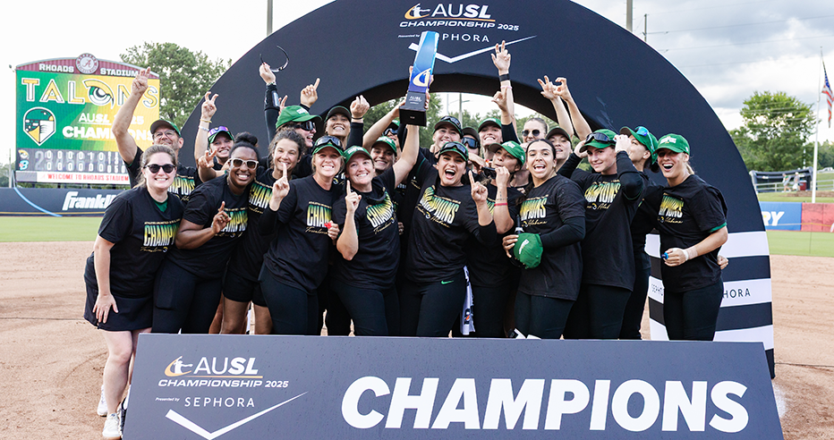 Members of the Talons, wearing their black t-shirts with "Champions" across the chest in green, yellow, and white lettering, and green hats, cheer as they raise the AUSL Championship trophy at Rhoads Stadium behind a Champions sign.