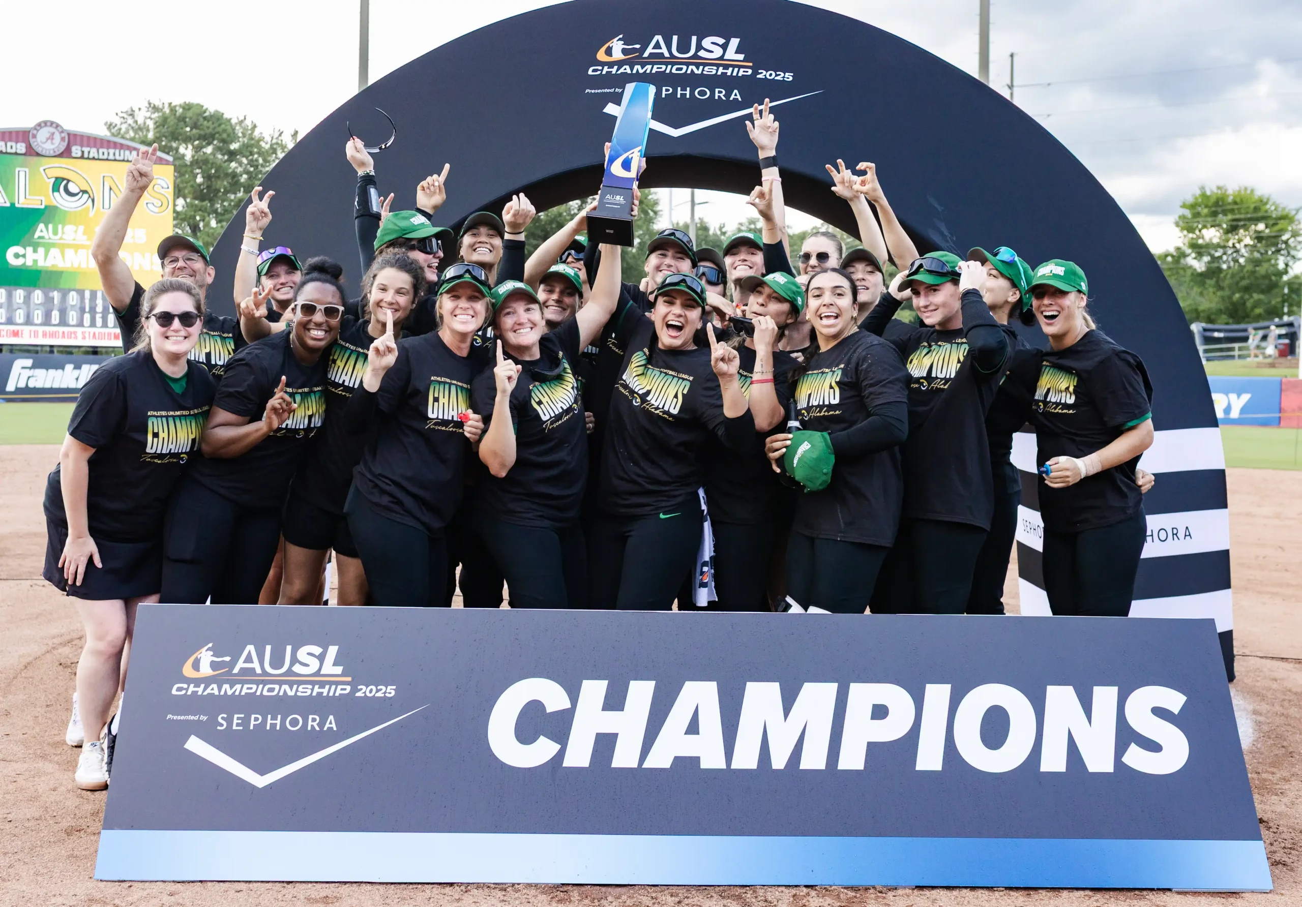 Members of the Talons, wearing their black t-shirts with "Champions" across the chest in green, yellow, and white lettering, and green hats, cheer as they raise the AUSL Championship trophy at Rhoads Stadium behind a Champions sign.